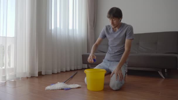 Ένας νεαρός πλένει το πάτωμα σε ένα διαμέρισμα. Βάζει υγρό πλύσης στον κουβά. Η έννοια της υπηρεσίας καθαρισμού. Έννοια ισότητας των φύλων. — Αρχείο Βίντεο