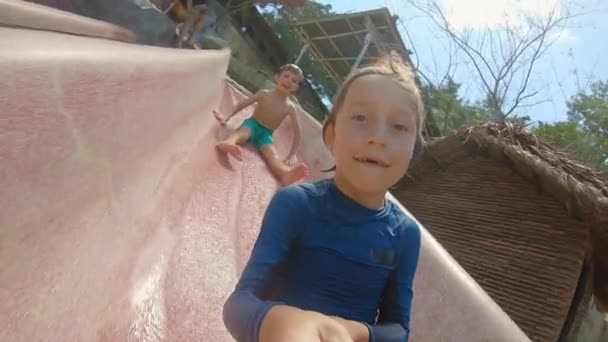 Två små pojkar glider ner för en rutschkana i en vattenpark. En av dem filmar en selfie video. — Stockvideo