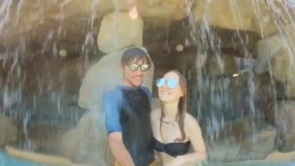 Una joven pareja tiene felices vacaciones en un complejo acuático. Ellos yacen bajo una cascada artificial en un centro de spa — Vídeos de Stock