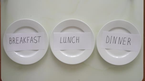 Empty plates with titles breakfast, lunch and dinner on a table. Woman replaces title dinner with the title fasting. Interval fasting concept. Skipping meals — Stock Video