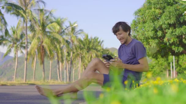Młody człowiek obsługuje drona siedzącego na trawie w tropikalnym otoczeniu. Koncepcja fotografii lotniczej lub wideo — Wideo stockowe
