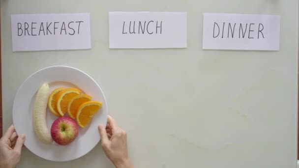 Las manos de una mujer ponen un plato con frutas frescas cortadas y arroz integral con verduras bajo los títulos desayuno y almuerzo respectivamente, después de lo cual bajo el título cena pone plato vacío con título — Vídeo de stock