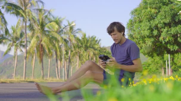 Egy fiatal férfi működtet egy drónt, aki egy füvön ül egy trópusi környezetben. Légi fényképezés vagy videofelvétel koncepció — Stock videók