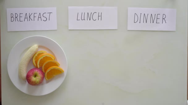 Las manos de una mujer ponen un plato con frutas frescas cortadas bajo el título desayuno y bajo el título almuerzo y cena pone platos vacíos con el título ayuno. Concepto de ayuno de intervalo. Saltarse las comidas — Vídeos de Stock