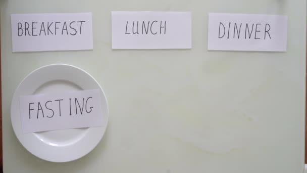 Pod tytułowym lunchem wkłada talerz z brązowym ryżem i warzywami, a pod tytułowym śniadaniem i kolacją wkłada puste talerze z tytułami postu. Koncepcja postu interwałowego. Pomijanie — Wideo stockowe