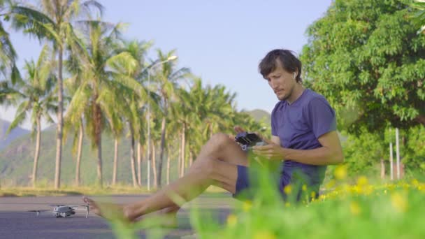Молодий чоловік керує безпілотником, який сидить на траві в тропічному оточенні. Дрон злітає. Концепція аерофотозйомки або відеозйомки — стокове відео