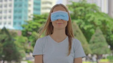 Genç bir kadın sokakta duruyor. Tıbbi bir yüz maskesi yanlış takıyor. Maskesi ağzını kapatmıyor, onun yerine gözlerini kapatıyor. Yüz maskesi kullanmanın yanlış yolu