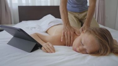 Yatak odasında genç bir çift var. Erkek, bir kadın için vücut masajı yapmayı bir tabletten online eğitim videosu izleyerek öğreniyor. Çevrimiçi eğitim kavramı. Covid yüzünden evde kalıyorlar.