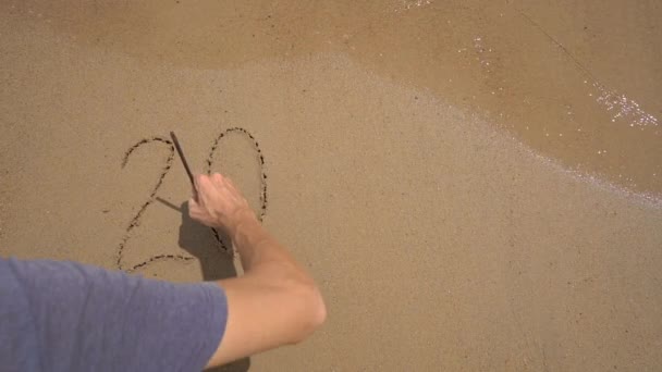 スローモーション撮影。男はビーチで砂の上に2020を書いています。人生の変化の概念。時代の変化 — ストック動画