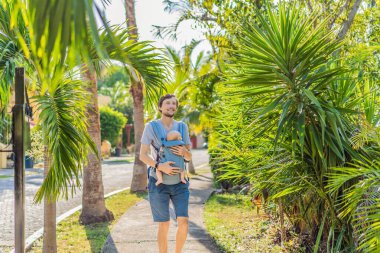 Genç bir baba, bebeğiyle birlikte doğada ebeveynlere bakan ergonomik bir bebek taşıyıcısının içinde dolaşıyor. Barışçıl bir ortamda bağlılık, sıcaklık ve modern ebeveynlik kavramı.
