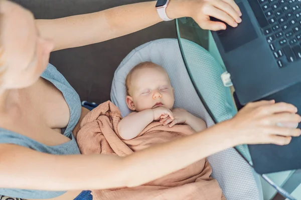Yeni doğmuş bebeği kucağında rahat bir bebek yuvasında huzur içinde uyurken aynı işi yapan anne evde dizüstü bilgisayarla çalışıyor. Uzaktan çalışma, annelik, aile ve kariyer dengeleme kavramı.