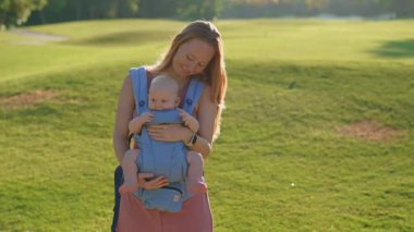 Genç anne, güneşli bir günde bebeğiyle parka gidip ergonomik bir taşıyıcıyla kaliteli zaman ve doğanın tadını çıkarıyor..