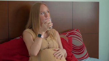 Hamile bir kadın yatağında otururken bir bardak su içer. Susuzluk, annelik sağlığı ve doğum öncesi bakım kavramı. Hamilelik süresince sağlıklı alışkanlıkları ve kendine bakımı teşvik etmek..