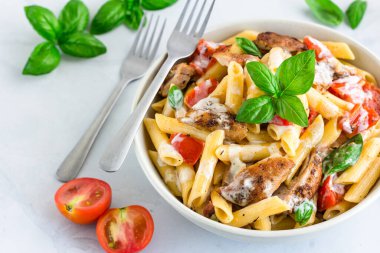 Beyaz soslu, tavuklu makarna, beyaz arka planda fesleğenle süslenmiş domates.