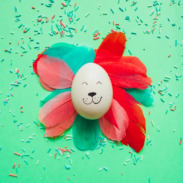 Bunny rabbit smiling face made of painted egg lay with colorful feathers on bright background. Easter minimal concept.