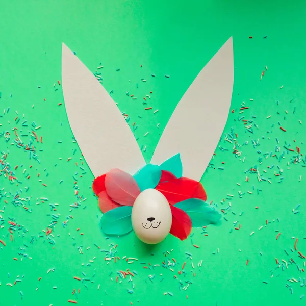 Bunny rabbit face and ears made of paper and painted egg lay on bright background. Easter minimal concept.