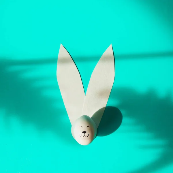 Easter egg with bunny rabbit face and paper ears on turquoise background with shadows of palm leaves.