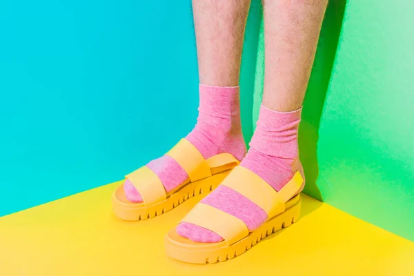 Male hairy legs in socks staying in women\'s sandals on bold background in the corner with strong shadows. Minimal pride concept. Body part