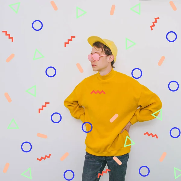 handsome man model in pink painted sunglasses, yellow baseball cap and oversize pullover among memphis geometry figures. crazy surreal art