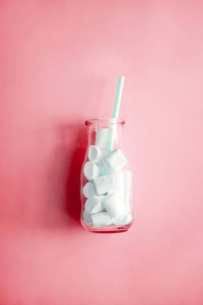 Creative minimalism still life on pastel pink colored background. Glass bottle with marshmallows and pink straw. Copyspace