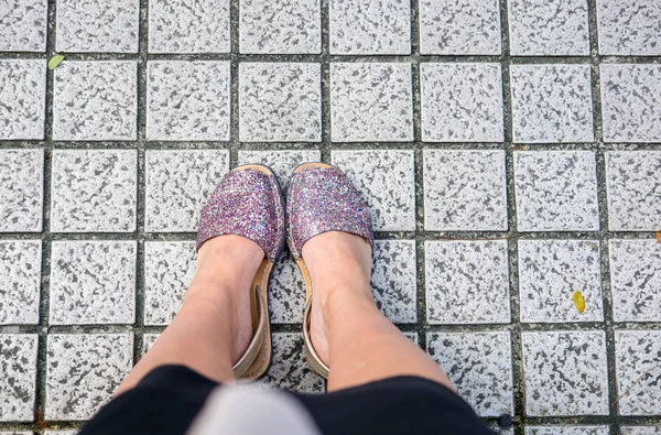 Overhead woman\'s feet in bright purple silver glitter sandals on grey square textured ground. Beauty, fashion minimalist blog concept. Copy space