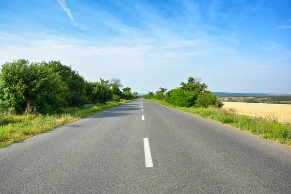 İki şeritli asfalt ülke road, ufuk ötesine gidiyor. Peyzaj manzaralı sigara kentsel driveway, bir buğday alanı, ağaçlar ve mavi gökyüzü beyaz bulutlar ile.
