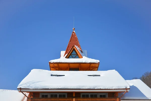 Snow on the roof of wooden house. Attic window of triangular shape on snow-covered roof on background of bright blue sky. Sunny winter day.