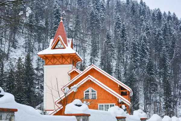 Winter landscape. Wooden cottage covered with snow against background of mountainside with spruce forest. Snow on the roof of wooden house.