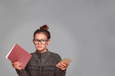 Asyalı kadın elinde bir kitap tutuyor ve telefonunu dikkatle Kömür Gri arka planda tutuyor..