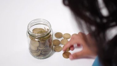 Genç bir kız, tasarruf, mali planlama ve erken yaştan itibaren öğrenme sorumluluğunu simgeleyen şeffaf cam bir kavanoza madeni para koyuyor.