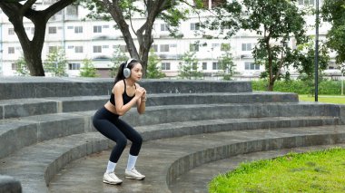 Genç Asyalı bir kadın parkta kulaklık takıyor, spor yapıyor, dışarıda egzersiz yapıyor, sağlık, esneklik, canlılık ve doğada dengeli ve sağlıklı bir yaşam tarzını teşvik ediyor...