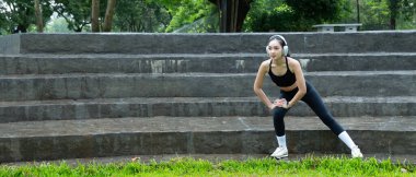 Genç Asyalı bir kadın parkta kulaklık takıyor, spor yapıyor, dışarıda egzersiz yapıyor, sağlık, esneklik, canlılık ve doğada dengeli ve sağlıklı bir yaşam tarzını teşvik ediyor...