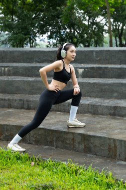 Genç Asyalı bir kadın parkta kulaklık takıyor, spor yapıyor, dışarıda egzersiz yapıyor, sağlık, esneklik, canlılık ve doğada dengeli ve sağlıklı bir yaşam tarzını teşvik ediyor...