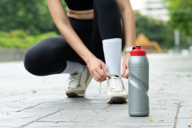 Kadın koşucu ayakkabı bağcıklarını bağlıyor. Bir şişe suyla parkta koşmaya hazırlanıyor..