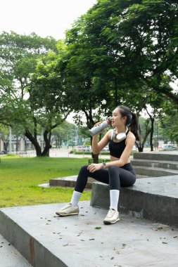 Bir kadın parkta bir bankta oturuyor, su şişesinden içiyor. Kulaklık takıyor ve dışarıda eğleniyor..