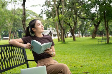 Genç Asyalı bir kadın parkta bankta oturmuş kitap okuyor ve güneşli günün tadını çıkarıyor..