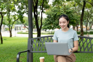 Parktaki bir bankta elinde kahve fincanıyla dizüstü bilgisayar kullanan genç bir kadın..