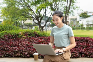 Parktaki bir bankta elinde kahve fincanıyla dizüstü bilgisayar kullanan genç bir kadın..