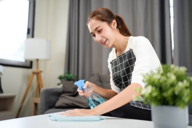 Bir kadın sprey şişesiyle bir masayı temizliyor. Gülümsüyor ve işini seviyor gibi görünüyor.