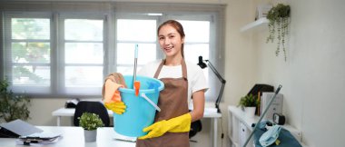 Ofiste dururken elinde temizlik aletleri ve ekipmanlarıyla kova tutan genç bir kadın..