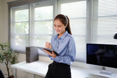 Ofisteki pencerenin önünde elinde tablet olan bir kadın gülümsüyor. Mavi gömlek ve siyah pantolon giyiyor.