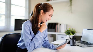 Bir kadın elinde tabletle bir masada oturuyor. Gülümsüyor ve işinden zevk alıyor. Tablet bir belgeye açık ve masanın etrafına dağılmış birkaç kağıt var..