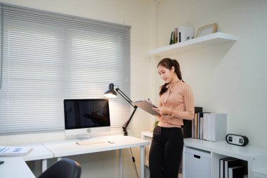 Bir kadın elinde bilgisayar monitörü ve bir panoyla masanın önünde duruyor.