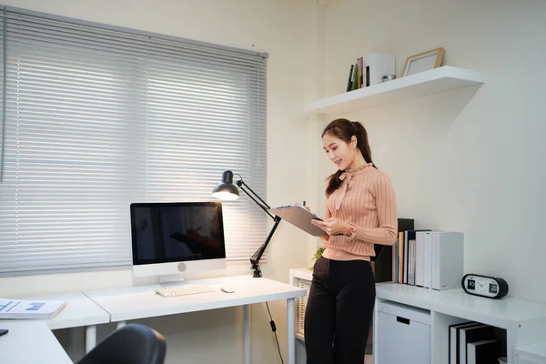 Bir kadın elinde bilgisayar monitörü ve bir panoyla masanın önünde duruyor.