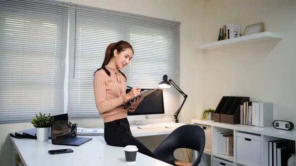 Bir kadın elinde bilgisayar monitörü ve bir panoyla masanın önünde duruyor..