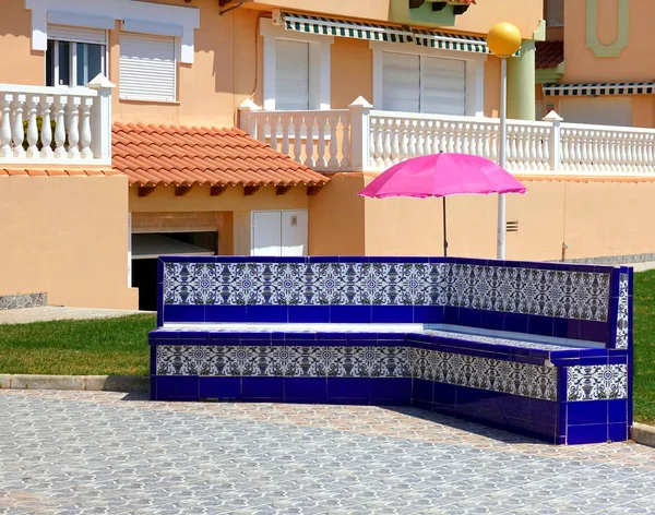 Terra cotta kum renkli apartman binası, süslü kiremitli mavi koltuk önünde karşı pembe güneş şemsiyesi tezat