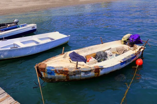 Parlak mavi bir denizde ahşap bir iskeleye demirlemiş küçük tekneler. Bir tanesi çok eski ve paslı metal plakalarla kaplanmış.