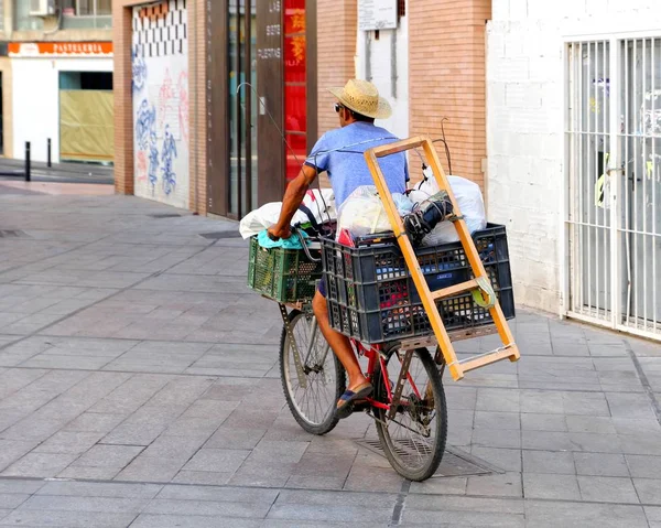 Murcia, İspanya - 4 Ağustos 2018: Hasır şapkalı bir adam, çeşitli kutu ve mallarla dolu bir İspanyol caddesinde bisiklet sürüyor