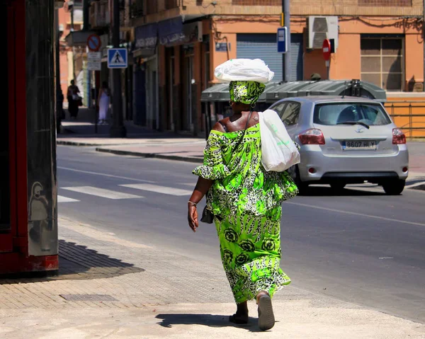 Cartagena, Murcia, İspanya - 01 Ağustos 2018: Afro-Karayipli bir kadın, parlak, yeşil, geleneksel Afrikalı giysiler içinde güneşli bir İspanyol caddesinde kafasında mallar taşıyor.