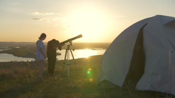 Девушка выходит из палатки к друзьям и смотрит в телескоп на закате лета — стоковое видео